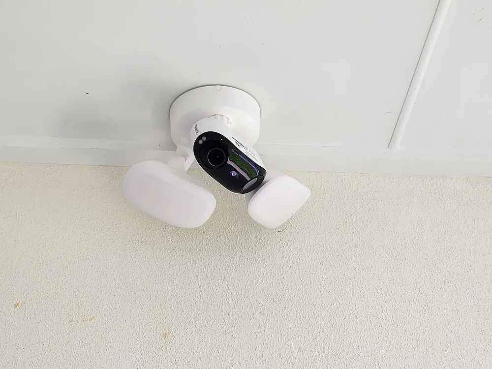 White floodlight security camera with twin lamps mounted under a porch ceiling